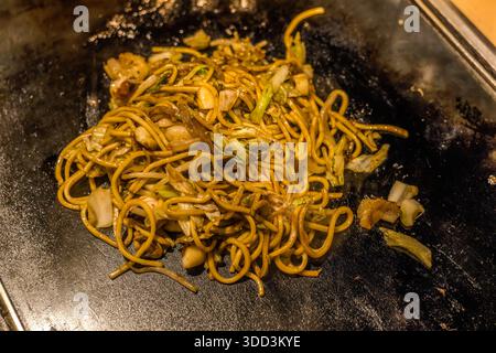 Gebratene Yakisoba-Nudeln liegen bereit auf der Teppanyaki-Grillplatte eines Restaurants in Osaka, Japan, gemischt mit sichtbaren Stücken Kohl, Zwiebel und Fleisch. Die gebratenen Weizen-Nudeln gehören neben Okonomiyaki zu den beliebtesten Grillgerichten von Osaka. Stockfoto