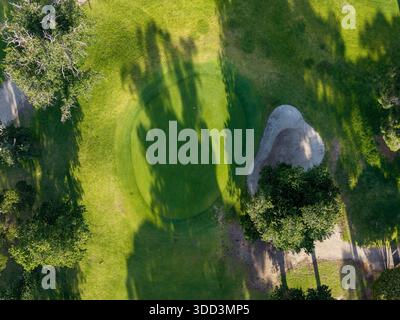 Ein Blick aus der Vogelperspektive zeigt ein perfekt gepflegtes Grün des Golfplatzes und einen angrenzenden Sandbunker, umgeben von üppigem Gras mit langen Baumschatten. Stockfoto