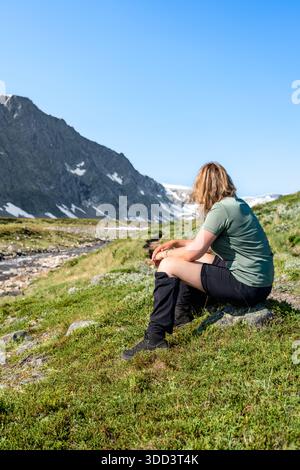 Frau sitzt neben einem Fluss und Wanderweg in den Trollheimen Bergen in Norwegen. Sie genießt die Landschaft im Freien und macht eine Pause in der Sommersonne Stockfoto