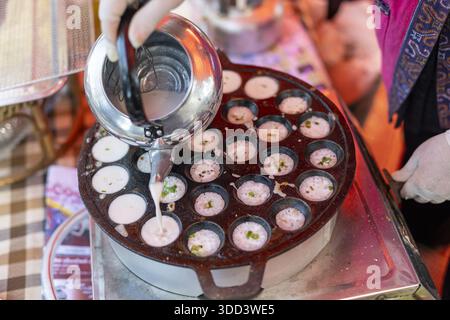 Küchenchef gießt süßen weißen Kokosnussmilchteig von Hand in eine heiße Metallpfanne und macht das traditionelle thailändische Dessert Kanom krok mit Sorgfalt und Aufmerksamkeit zu einem Feinkost Stockfoto
