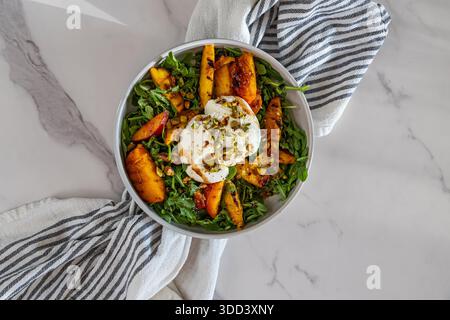 Ein Blick von oben auf einen lebhaften Sommersalat mit gegrillten Pfirsichen, cremiger Burrata und Rucola, garniert mit gehackten Pistazien. Stockfoto