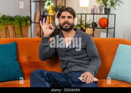 Ein indischer Mann zu Hause trägt kabellose Kopfhörer spielt Naturgeräusche, Atmen, Meditieren und Lächeln. Ein Araber auf dem Sofa hört ruhige Geräuschshows Stockfoto