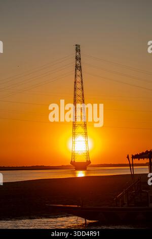 Sonnenuntergang über dem Brahmaputra River, wo ein Hochspannungsturm vor dem glühenden Himmel steht und Industrie mit Natur verbindet. Stockfoto