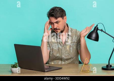 Ein Mann mittleren Alters sitzt am Tisch und erhält schlechte Nachrichten. Er erlebt eine Niederlage bei Online-Spielen auf einem Laptop und zeigt eine deutliche Enttäuschung. Junger Erwachsener, isoliert auf blauem Hintergrund, der sich verärgert fühlt Stockfoto
