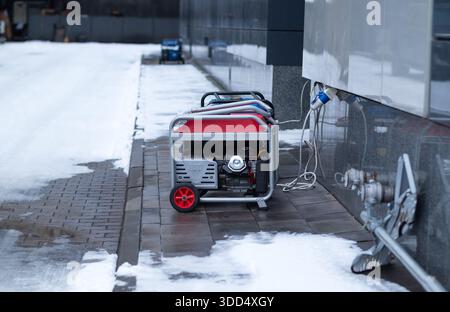 Tragbarer roter Benzingenerator zur Notstromversorgung eines modernen Gebäudes an einem verschneiten Wintertag Stockfoto