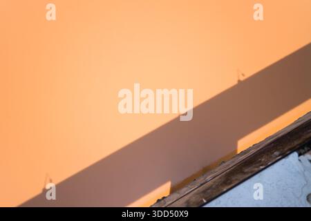 Der diagonale Schatten fällt über eine glatte pfirsichfarbene Wand, die von einem verwitterten Holzrand und einer strukturierten weißen Oberfläche durchzogen wird, wodurch eine minimalistische abstrakte Komposition in natürlichem Licht entsteht. Stockfoto