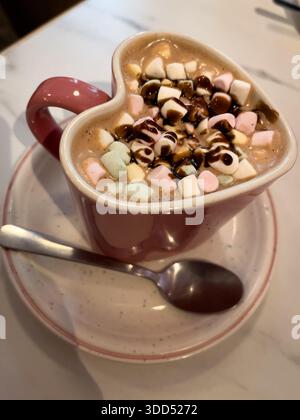 Eine herzförmige Kaffeetasse mit heißer Schokolade mit Marshmallows und Schokolade darüber. Der Becher ist auf einem weißen Teller mit einem Löffel daneben. Kriegsbegriff Stockfoto