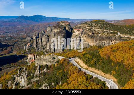 Die geschwungene Bergstraße führt durch den lebhaften Herbstwald zu den historischen Meteora-Klöstern, die sich auf hoch aufragenden Felsformationen thronen, mit einem atemberaubenden Blick auf die Thessalien-Landschaft und die fernen Berge unter einem klaren blauen Himmel. Stockfoto