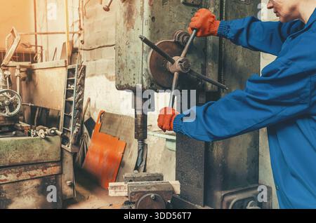 Fabrikarbeiterin in blauer Arbeitskleidung und orangefarbenen Sicherheitshandschuhen, die industrielle Bohrmaschine in der Fertigungswerkstatt bedienen. Concep Stockfoto