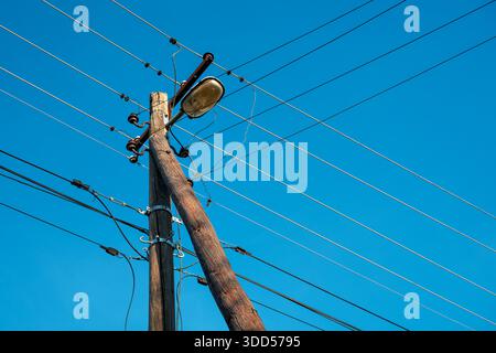 Verwitterter hölzerner Strommast mit verworrenen Stromkabeln, Keramikisolatoren und einer alten Straßenlampe vor einem klaren, hellblauen Himmel. Wählen Sie Stockfoto