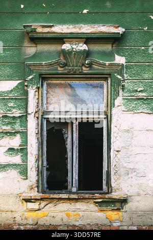 Verwittertes Vintage-Fenster mit abblätternder grüner Farbe, gebrochenem Glas und zerbröckelnder Fassade in einem alten verlassenen Gebäude. Vertikales Bild. Stockfoto