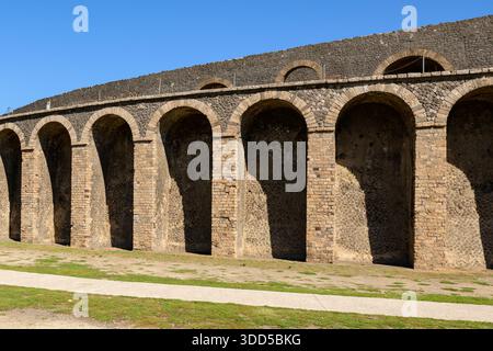 Eine Reihe von antiken Steinbögen bildet die Außenmauer des römischen Amphitheaters in Pompeji, Italien. Helles Sonnenlicht hebt das strukturierte vulkanische Mauerwerk und die geometrischen Linien unter einem klaren blauen Himmel hervor und unterstreicht die historische Architektur. Stockfoto