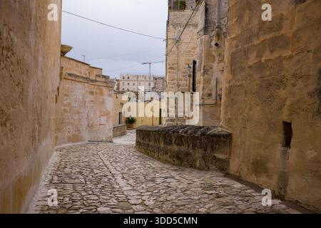 Eine enge Kopfsteinpflasterallee kurvt sich zwischen verwitterten Steinhäusern in Matera, Italien. Gedämpftes Tageslicht unterstreicht strukturierte Wände und den zeitlosen Charakter der Altstadt. Stockfoto