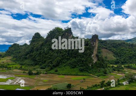 Steile Kalksteinformationen, bedeckt mit dichtem grünen Waldturm über Flickentrickfeldern in der ländlichen Landschaft zwischen Vang Vieng und Phou Khoun, Laos. Die Szene zeigt dramatische vertikale Klippen, überflutete Felder und einen Himmel voller texturierter Wolken und blauer Flecken. Stockfoto
