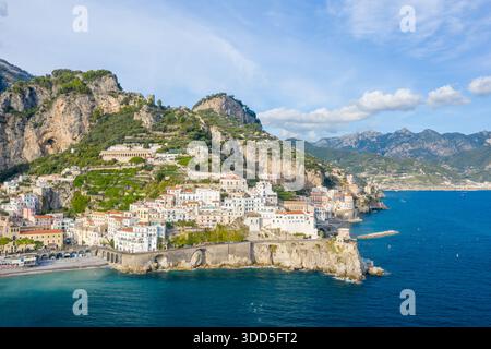 Eine weite Luftperspektive fängt die weiß getünchten Gebäude von Amalfi, Italien, ein, die zerklüftete Klippen hinunterragen und zum leuchtend blauen Mittelmeer führen. Die dramatische Küste bietet historische Steinbögen, üppiges Grün und eine bergige Kulisse unter klarem Himmel. Stockfoto