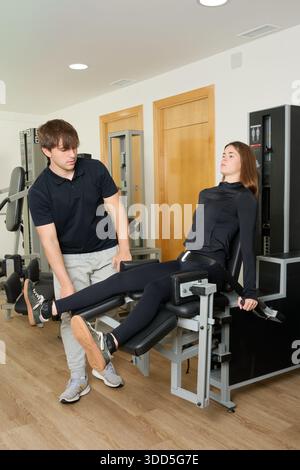 Junge Frau trainiert Beine auf einem Fitnessgerät mit professioneller persönlicher Trainerführung und Unterstützung der Muskelkraft Stockfoto