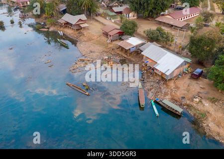 Aus der Vogelperspektive sehen Sie rustikale hölzerne Docks und Longtail-Boote entlang des Ufers in der Region der 4000 Inseln im Süden von Laos. Traditionelle Pfahlhäuser, Palmen und ruhiges blaues Wasser schaffen eine ruhige Umgebung am Fluss mit sichtbarem Dorfleben und natürlichen Texturen. Stockfoto