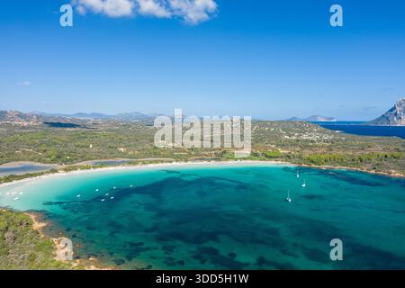 Blick aus einem Hochwinkel auf den Strand Cala Brandinchi auf Sardinien mit einem weiten Bogen aus türkisfarbenem Wasser, ankerbaren Booten und einer üppigen grünen Küste unter einem klaren Himmel mit weit entfernten Bergen. Stockfoto