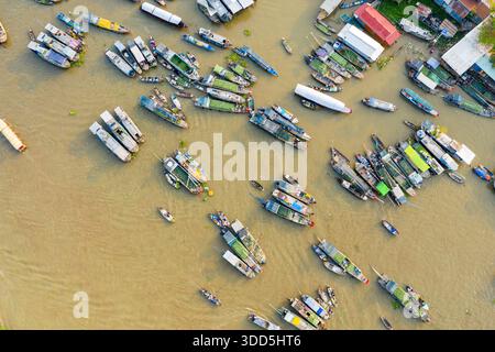 Die direkte Ansicht von oben zeigt ein lebhaftes Muster von Marktbooten, die sich auf dem Fluss am schwimmenden Markt Cai Rang in Can Tho, Mekong Delta, Vietnam versammelt haben. Das Bild zeigt überdachte Schiffe, grüne Produkte und Gebäude am Fluss unter klarem Tageslicht, die den pulsierenden Handelsfluss einfangen. Stockfoto