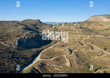 Aus der Vogelperspektive eine serpentine Landstraße, die sich durch trockene, felsige Hügel in der Nähe von Nafplio, Griechenland, schlängelt. Die sonnendurchflutete Landschaft zeichnet sich durch trockene Texturen, karge Vegetation und dramatische natürliche Formationen unter einem klaren blauen Himmel aus. Stockfoto