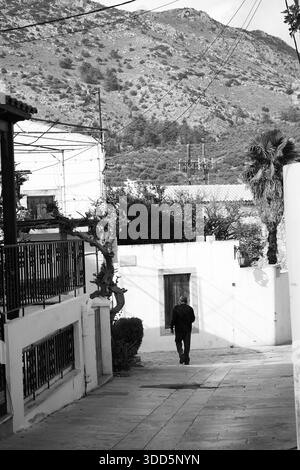 Vertikale Schwarzweißaufnahme eines älteren Mannes, der eine ruhige Steingasse in Archanes, Kreta, in Griechenland, in Richtung einer malerischen Bergkulisse hinuntergeht. Stockfoto