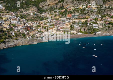 Die weite Luftperspektive von Positano, Italien, zeigt lebendige Reihenhäuser, die steile Klippen hinunter zu einem von Sonnenschirmen gesäumten Sandstrand führen. Das tiefblaue Mittelmeer ist mit kleinen Booten gesäumt, während üppiges Grün und dramatische Kalksteinhänge die berühmte Amalfiküste umrahmen. Stockfoto