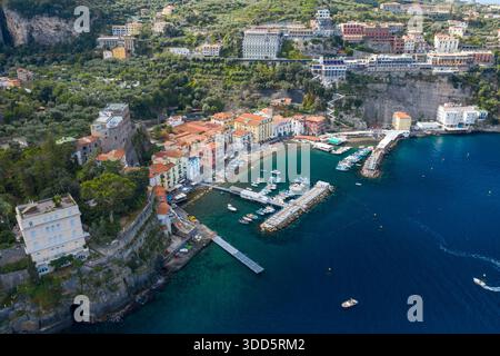 Hochwinkelpanorama von Sorrent, Italien, mit lebhaften Häusern um einen kleinen Yachthafen und dramatischen Klippen, die sich dahinter erheben. Die Szene bietet türkisfarbenes mediterranes Wasser, üppiges Grün und eine lebhafte Küstenatmosphäre. Stockfoto