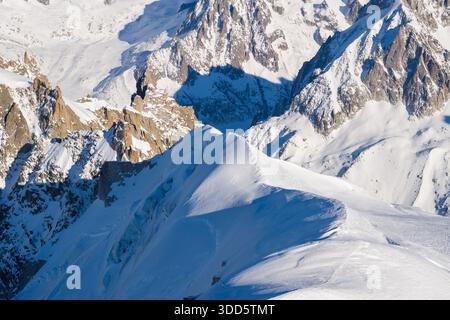 Im Vordergrund erhebt sich ein scharfer, unberührter Schneegrat, der sich vor dramatischen, sonnendurchfluteten Granitgipfeln und tiefblauen Schatten im Mont Blanc-Massiv erhebt. Stockfoto