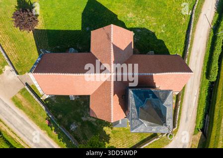 Direkte Drohnenaufnahme von oben nach unten eines kreuzförmigen, rot gekachelten Kirchendachs, umgeben von grünen Rasenflächen und Landstraßen in Cuncy les Varzy, Burgund, an einem sonnigen Sommertag. Stockfoto