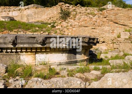Detaillierte Löwenkopfschnitzereien aus Stein zieren die alte kreisförmige Struktur des Nymphaeums von Herodes Atticus in Olympia, Griechenland. Warmes Sonnenlicht unterstreicht den verwitterten Marmor und das strukturierte Mauerwerk inmitten von grasbewachsenen Ruinen und historischen Ziegelmauern. Stockfoto