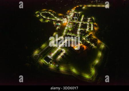 Drohnenperspektive, die ein kleines Dorf bei Nacht mit gewundenen Straßen und Wohngebäuden einfangen, die hell von Straßenlaternen beleuchtet werden. Die Szene zeigt geometrische Muster, kontrastierende Lichtquellen und das organisierte Layout der Siedlung, umgeben von Dunkelheit. Stockfoto
