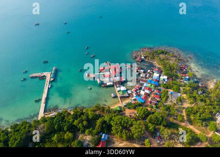 Die Aussicht über den Kopf zeigt ein farbenfrohes Fischerdorf mit lebhaften Dächern, Holzpiers und Booten, die in klarem türkisfarbenem Wasser am Coconut Beach, Koh Rong, Kambodscha, verankert sind. Üppiges tropisches Grün und felsige Küste umrahmen die ruhige Küstensiedlung. Stockfoto