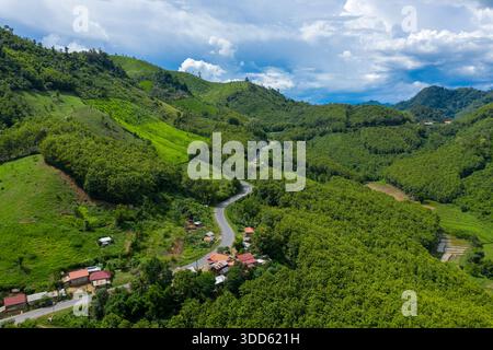 Kurvige Landstraßen schlängeln sich zwischen kleinen Häusern und dichten bewaldeten Hügeln im Norden von Laos. Üppiges Grün und dramatische Sommerwolken schaffen eine ruhige ländliche Landschaft mit lebendigen natürlichen Texturen. Stockfoto