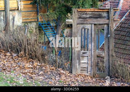 Eine Holztür steht in der Nähe eines verwitterten Gebäudes, umgeben von getrockneten Blättern und Gras. Blaue Treppen führen zum Gebäude. Die Einstellung zeigt Anzeichen von Stockfoto