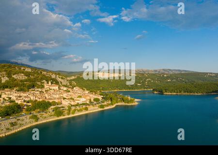 Blick aus der Vogelperspektive auf das Dorf Bauduen im warmen Sonnenlicht, eingebettet zwischen bewaldeten Hügeln und dem lebhaften türkisfarbenen Wasser des Lac de Sainte-Croix unter einem teilweise bewölkten Himmel. Stockfoto