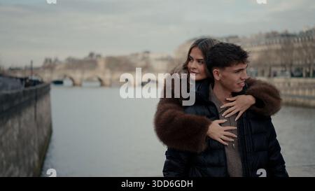 Ein romantisches Paar umarmt auf einer Brücke in Paris Frankreich mit der seine und der Stadtlandschaft im Hintergrund die junge Frau umarmt den Mann, der beide trägt Stockfoto