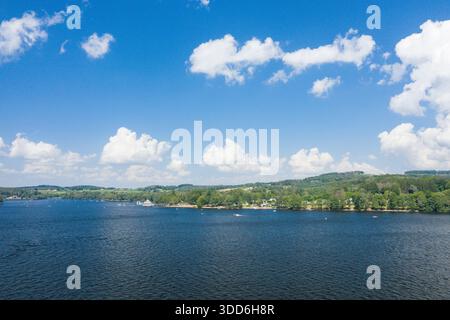 Eine weite Ausdehnung von tiefblauem Wasser erstreckt sich zu einer üppigen, von Bäumen gesäumten Küste mit sanften grünen Hügeln und verstreuten Booten unter einem hellen Himmel voller geschwollener weißer Wolken. Stockfoto