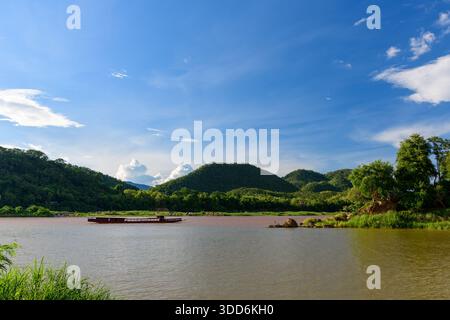 Ein breiter Abschnitt des schlammigen Mekong River fließt vorbei an grünen Hügeln und dichter Flusspflanze in der Nähe von Luang Prabang, Laos. Ein traditionelles Langboot driftet unter einem leuchtend blauen Himmel mit verstreuten Wolken auf dem Wasser und schafft eine lebendige natürliche Landschaft. Stockfoto