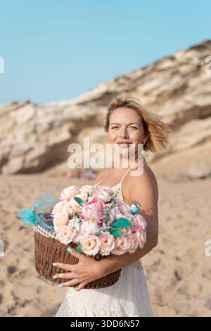 Lächelnde junge Frau im Kleid, die einen Korb mit rosa Blumen am Sandstrand hält und Sommerurlaub in der Nähe von felsigen Klippen genießt. Schöne Dame mit kurzen blonden Haaren, die in die Kamera schauen Stockfoto