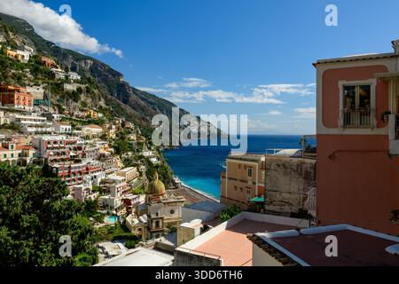 Helle pastellfarbene Gebäude kaskadieren den steilen Hügel von Positano, Italien, mit Blick auf das leuchtend blaue Wasser der Amalfiküste. Die Szene bietet farbenfrohe mediterrane Architektur, zerklüftete Klippen und einen klaren Himmel mit verstreuten Wolken. Stockfoto