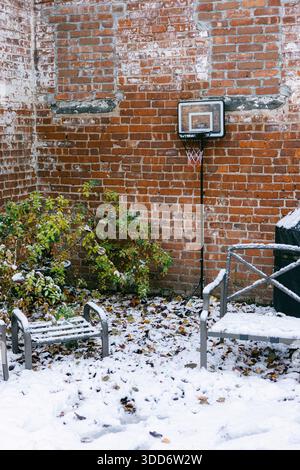 Schnee bedeckt den Boden in einem Innenhof mit Basketballkorb, leeren Stühlen und kleinen Pflanzen in der Nähe einer Backsteinmauer. Stockfoto