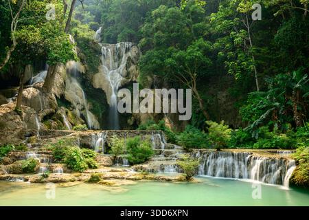 Gestufte Kalksteinkaskaden münden in türkisfarbene Pools inmitten des dichten tropischen Waldes an den Kuang Si Wasserfällen im Norden von Laos. Sonnenlicht durchströmt lebendiges grünes Laub und hebt die natürlichen Strukturen und die üppige Landschaft hervor. Stockfoto