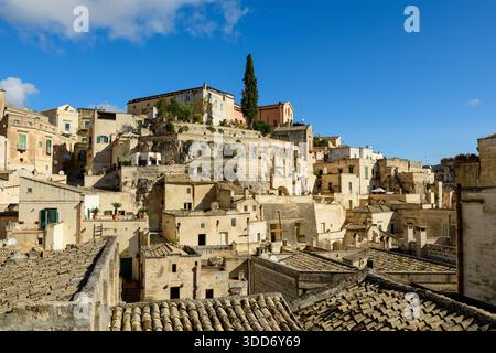 In Matera, Italien, erheben sich geschichtsträchtige Steinhäuser und Terrassendächer entlang des Hügels, die von hellem Sonnenlicht mit reichen Texturen und einem leuchtend blauen Himmel über dem Hügel getaucht sind. Stockfoto