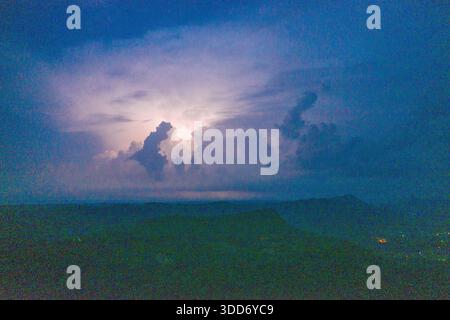 Große Gewitterwolken leuchten nachts mit innerem Blitz über einem dunklen Bergrücken. Die Szene zeigt dramatische Wolkenformationen, mehrschichtige Silhouetten und eine stimmungsvolle blaue Dämmerungsatmosphäre. Stockfoto