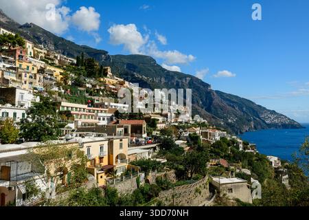 Farbenfrohe Terrassengebäude von Positano, Italien, kaskadieren steil abfallenden Hängen in Richtung des tiefblauen Mittelmeers. Üppiges Grün und zerklüftete Kalksteinberge umgeben das lebendige Küstendorf unter einem klaren Himmel mit verstreuten Wolken. Stockfoto