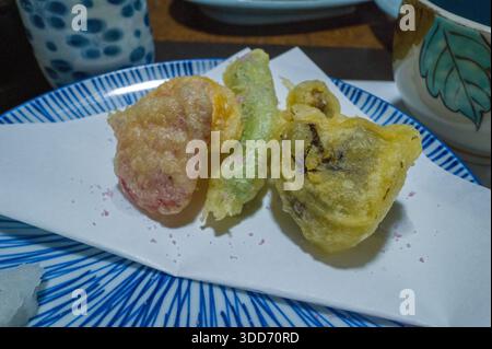 Drei verschiedene Gemüsetempura-Stücke sind auf weißem Papier auf einer blau gestreiften Keramikplatte angeordnet, umgeben von elegantem japanischem Geschirr. Die Nahaufnahme hebt den goldenen, luftigen Teig und die dezenten Farben in einem raffinierten Ambiente hervor. Stockfoto