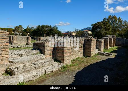 Verwitterte Steinfundamente und Ziegelsäulen stehen unter freiem Himmel an der archäologischen Stätte Paestum, Italien. Die Szene wird von hellem Sonnenlicht unter einem leuchtend blauen Himmel beleuchtet, umgeben von Grün und fernen Bergen. Stockfoto