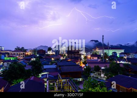 Heller Blitz beleuchtet den Abendhimmel über den Dächern von Vang Vieng, Laos, mit dramatischen Kalksteinbergen und treibendem Nebel im Hintergrund. Die Stadt leuchtet mit verstreuten Lichtern und unterstreicht den Kontrast zwischen urbanem Leben und wilder Landschaft. Stockfoto