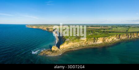 Panoramablick auf dramatische Klippen und felsige Küste in Pointe du hoc, Normandie, mit grünem Ackerland, das unter klarem Himmel auf das tiefblaue Meer trifft. Stockfoto