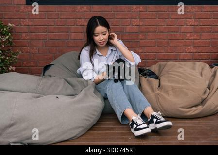 Junge asiatische Frau, die ein schwarzes Kätzchen hält, während sie sich auf Sitzsackstühlen entspannt Stockfoto